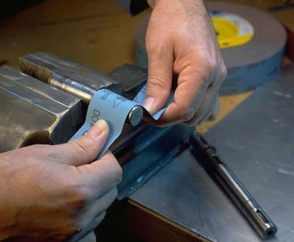 Hands using 600 grit emery cloth roll in a vise for precise sanding on metal surfaces.