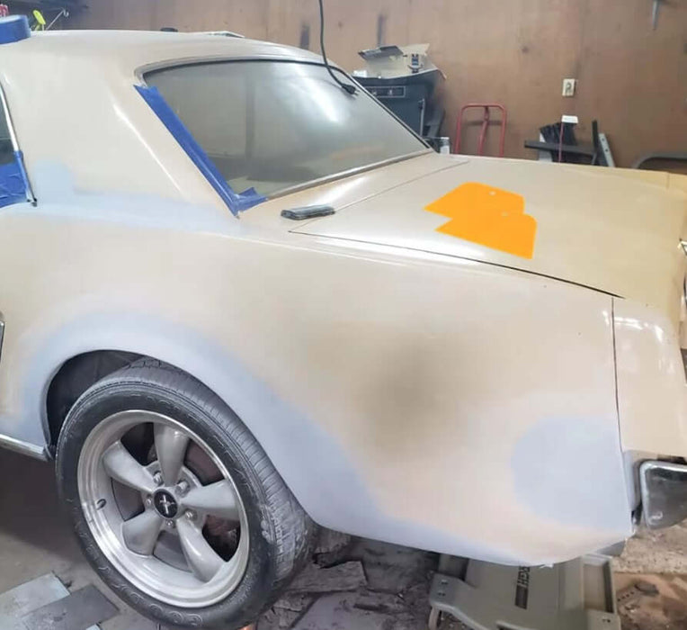 Classic car undergoing bodywork with automotive scraper spreader on the surface for filler application in garage setting.